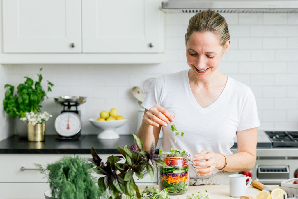 Holistic wellness coach placing salad greens into a glass jar, highlighting a kitchen setup for a brand shoot.