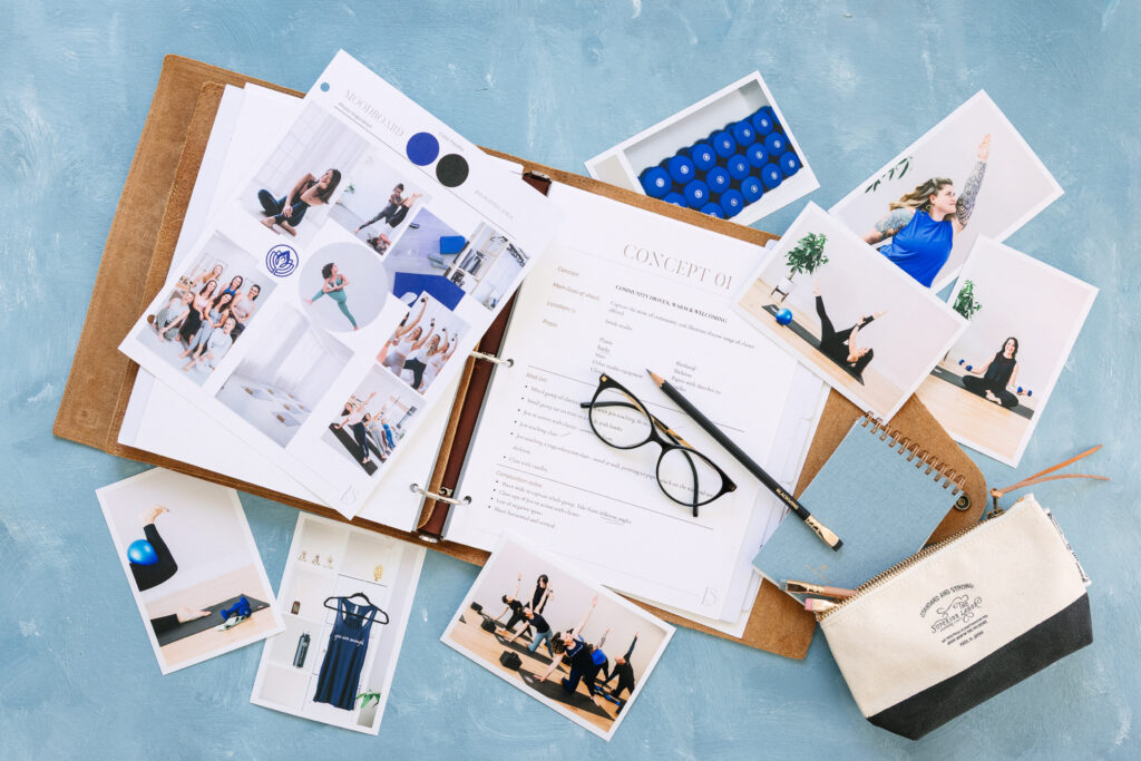 An open ring binder covered in photographs, a pair of black reading glasses, a pencil, and a pencil case on a blue painted styling mat.