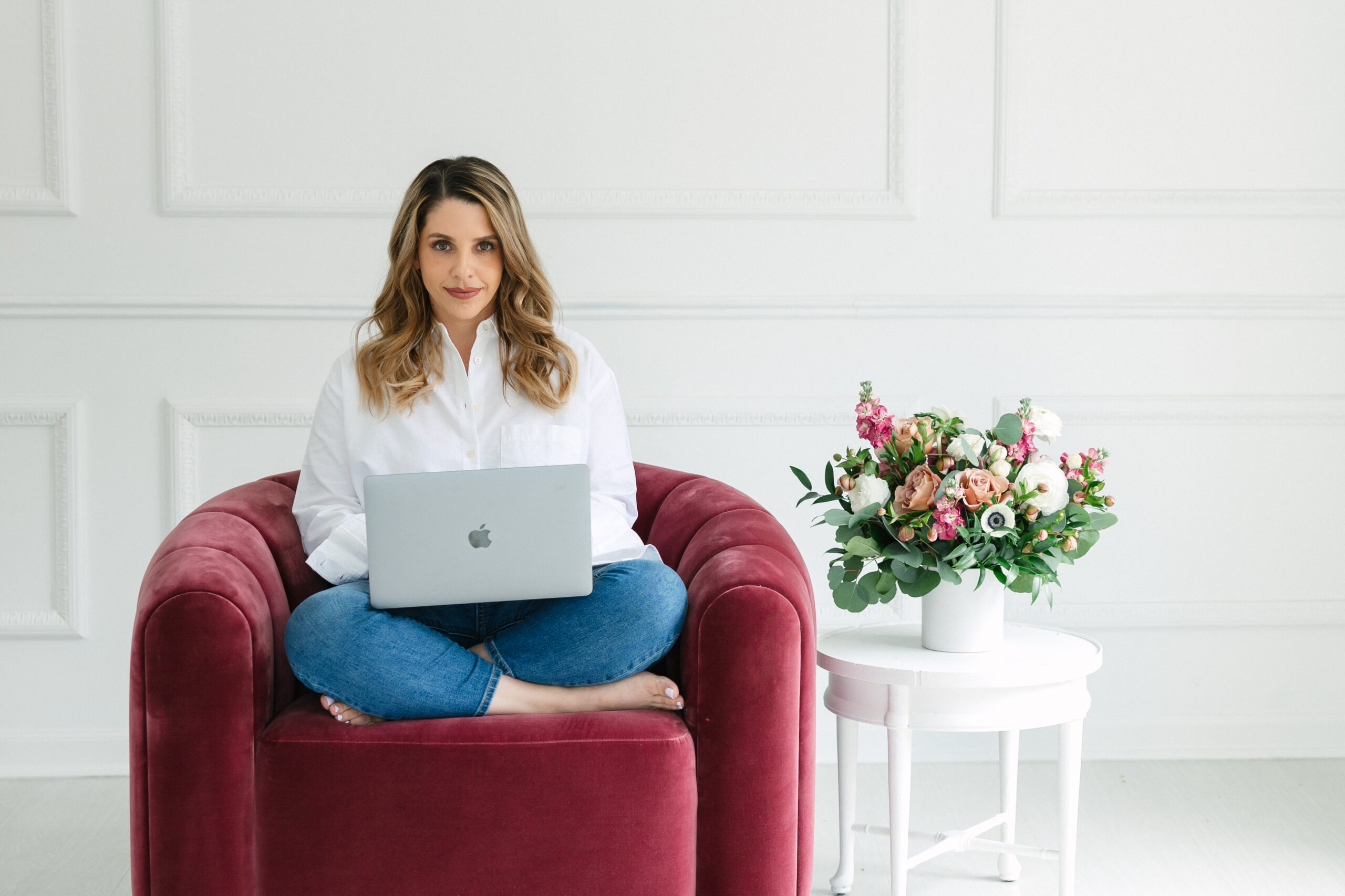 Woman sat crisscrossed in a rose colored velvet chair with a laptop.