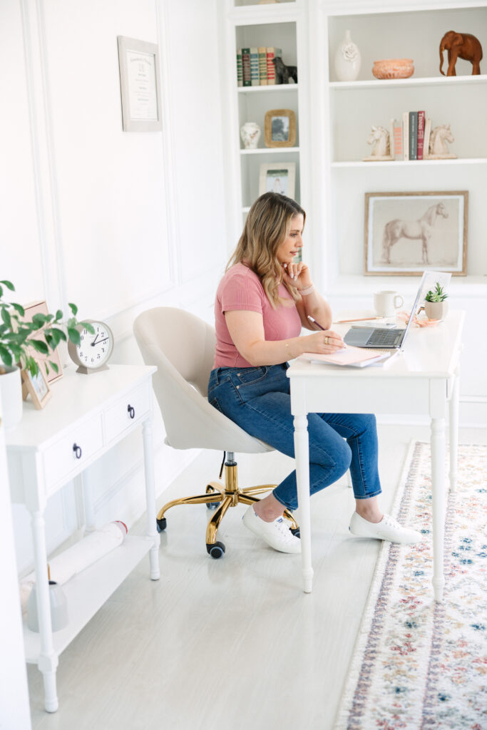 A woman wearing blue jeans and a rose colored tshirt sat at a desk, demonstrating a "The Connection" outfit during a brand photo shoot.