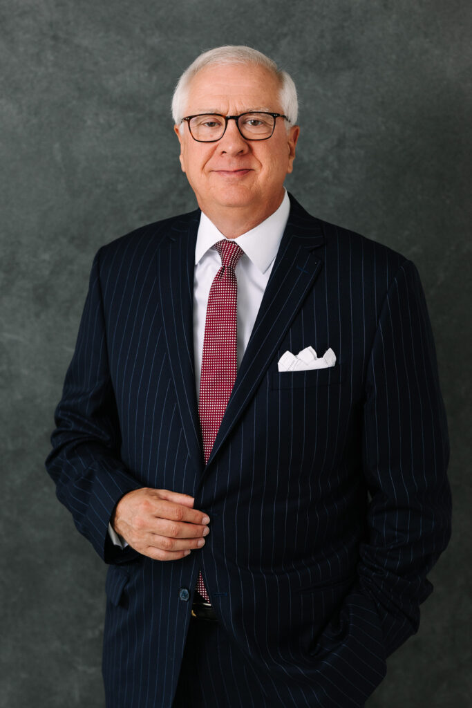 Professional headshot of a businessman wearing glasses, a dark suit, and a red tie against a gray studio background.